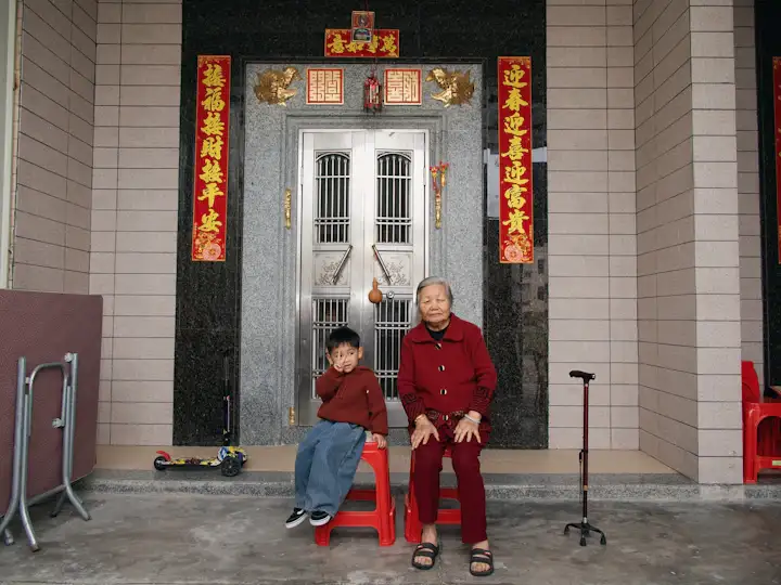 An older Asian woman with a cane sitting with her grandson at the front door of their home
