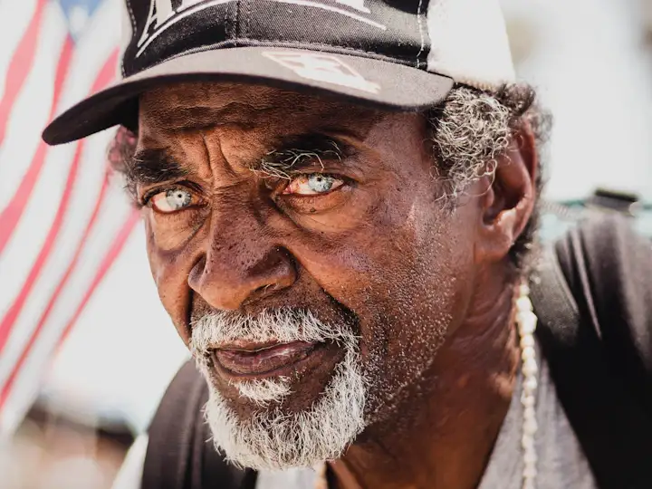 An older Black man in a cap in front of an American flag